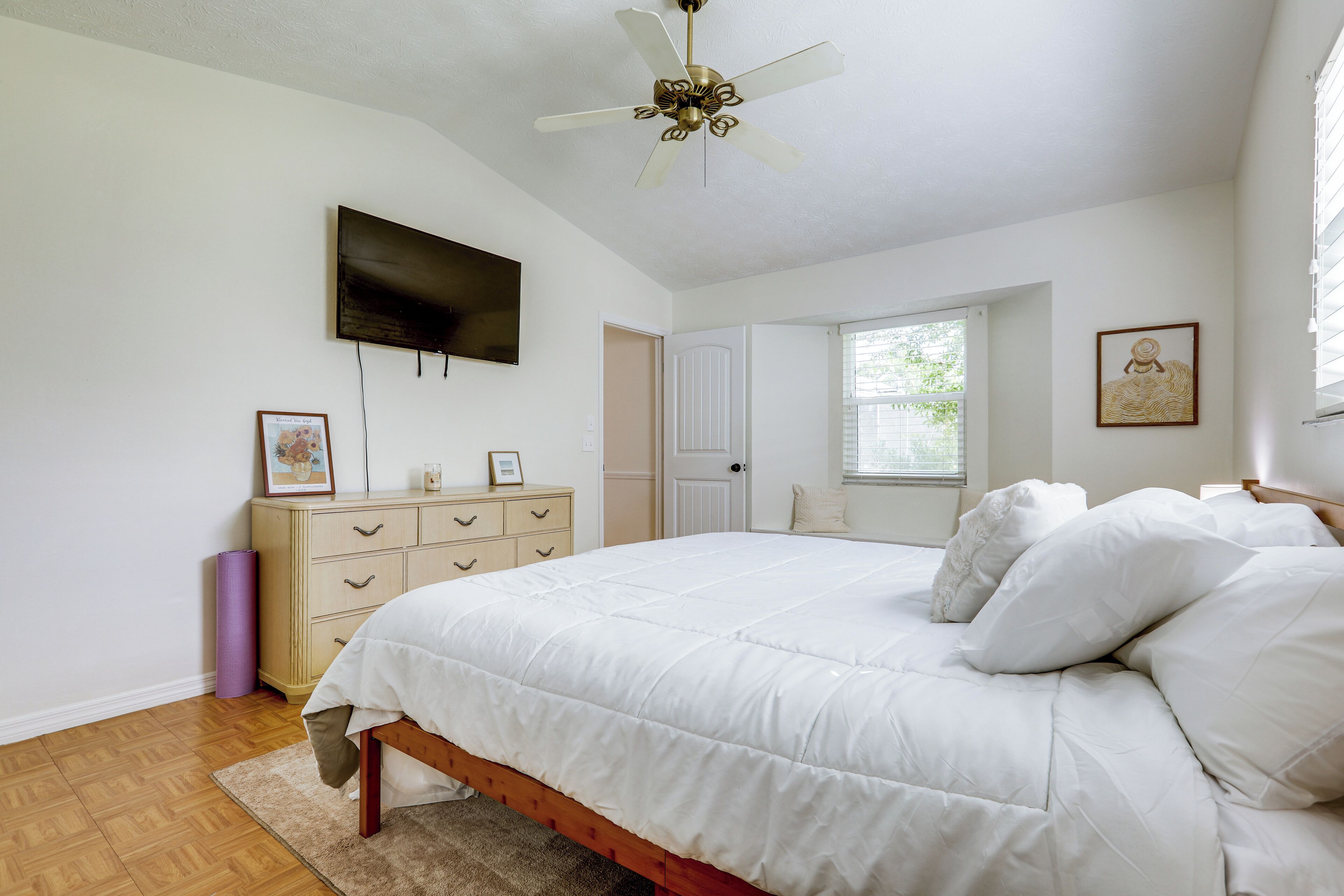 A bedroom with a bed, a ceiling fan, a TV, a dresser, a window, and a door.