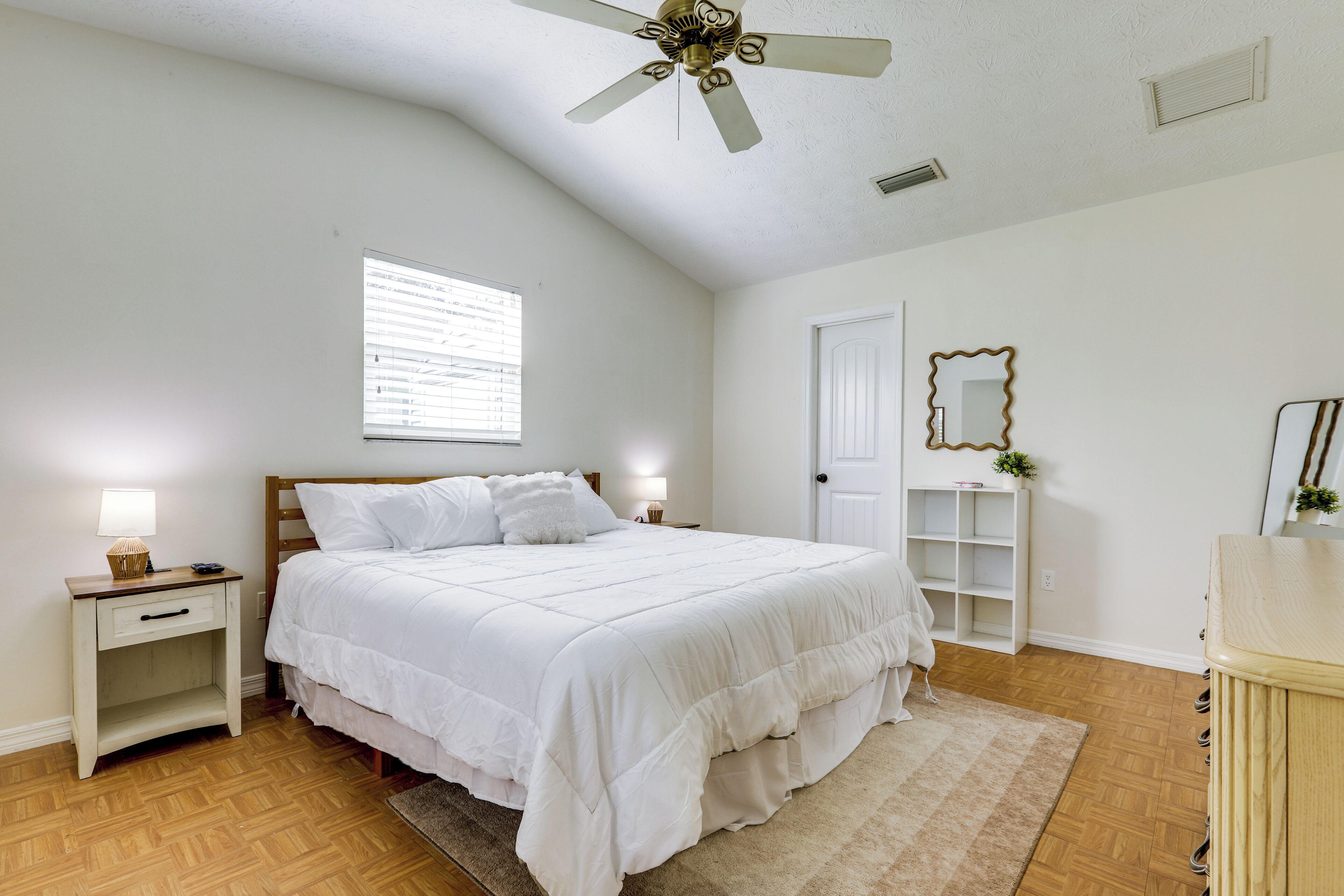 A bedroom with a bed, nightstands, a ceiling fan, a window, a door, and a mirror.