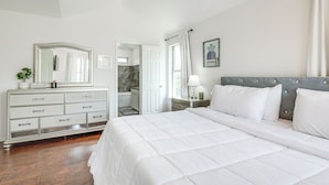 Habitación de hotel con una cama grande, cómoda, espejo, puerta que da a un baño y un cuadro en la pared.