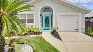 Una casa de un piso con puerta principal turquesa, un portón de garaje y un pequeño jardín delantero con plantas y una palmera.