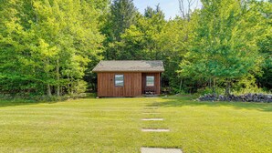 Une petite cabane en bois, dotée de deux fenêtres et d’une porte, entourée d’une cour verdoyante et d’arbres denses.