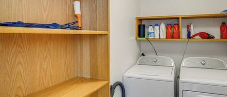 A laundry area with two white washing machines, wooden shelves with cleaning supplies, and a blue towel on the left shelf.