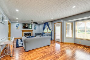 A spacious living room with hardwood flooring, a grey sofa, a fireplace, and large windows with blinds.