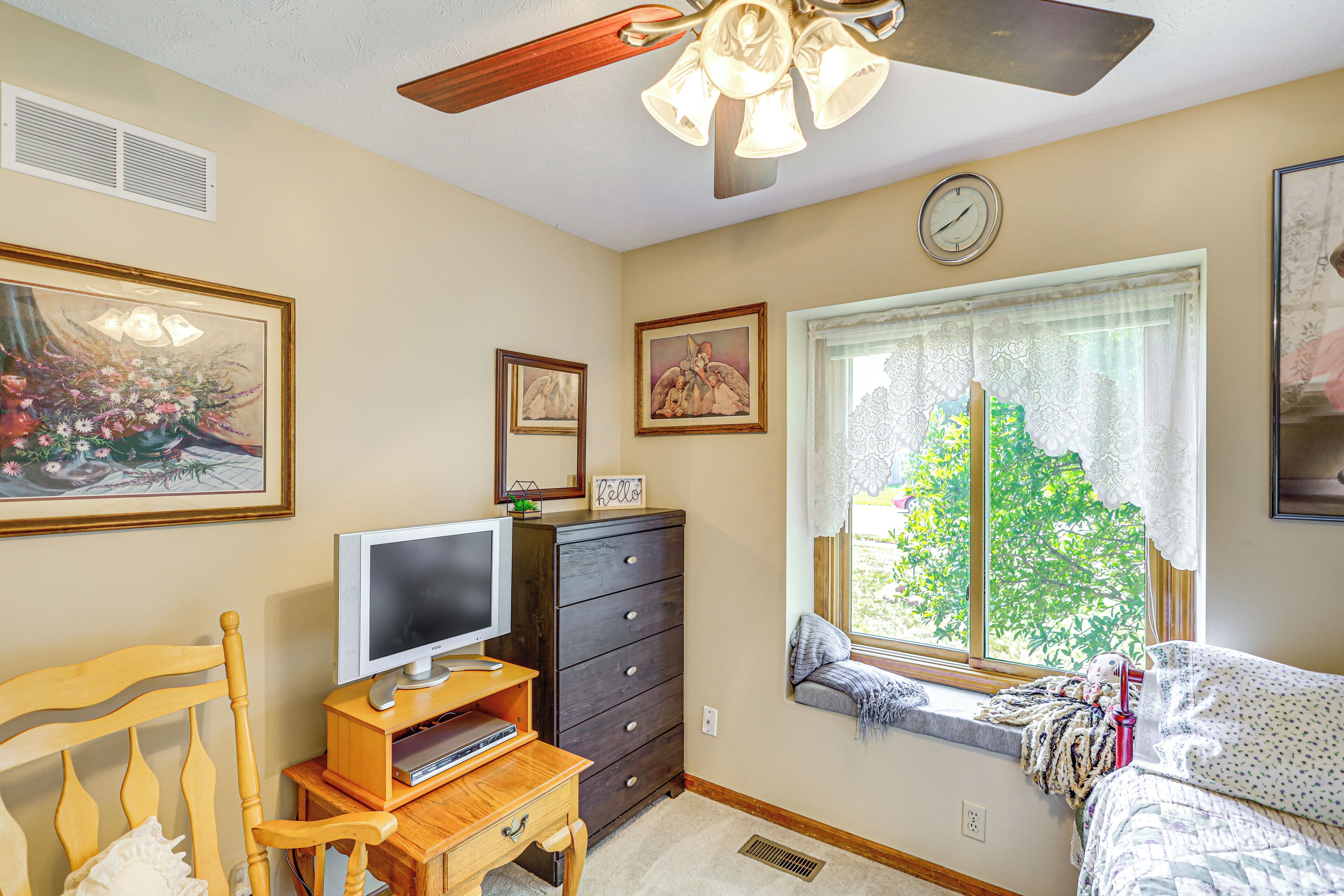 A bedroom with a bed, a dresser, a television on a stand, a window with a view of trees, and a ceiling fan.