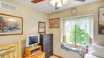 A bedroom with a bed, a dresser, a television on a stand, a window with a view of trees, and a ceiling fan.