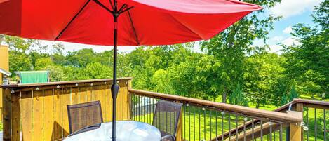 Una terraza de madera con una mesa y sillas de vidrio, un paraguas rojo y vistas a un césped verde y árboles.