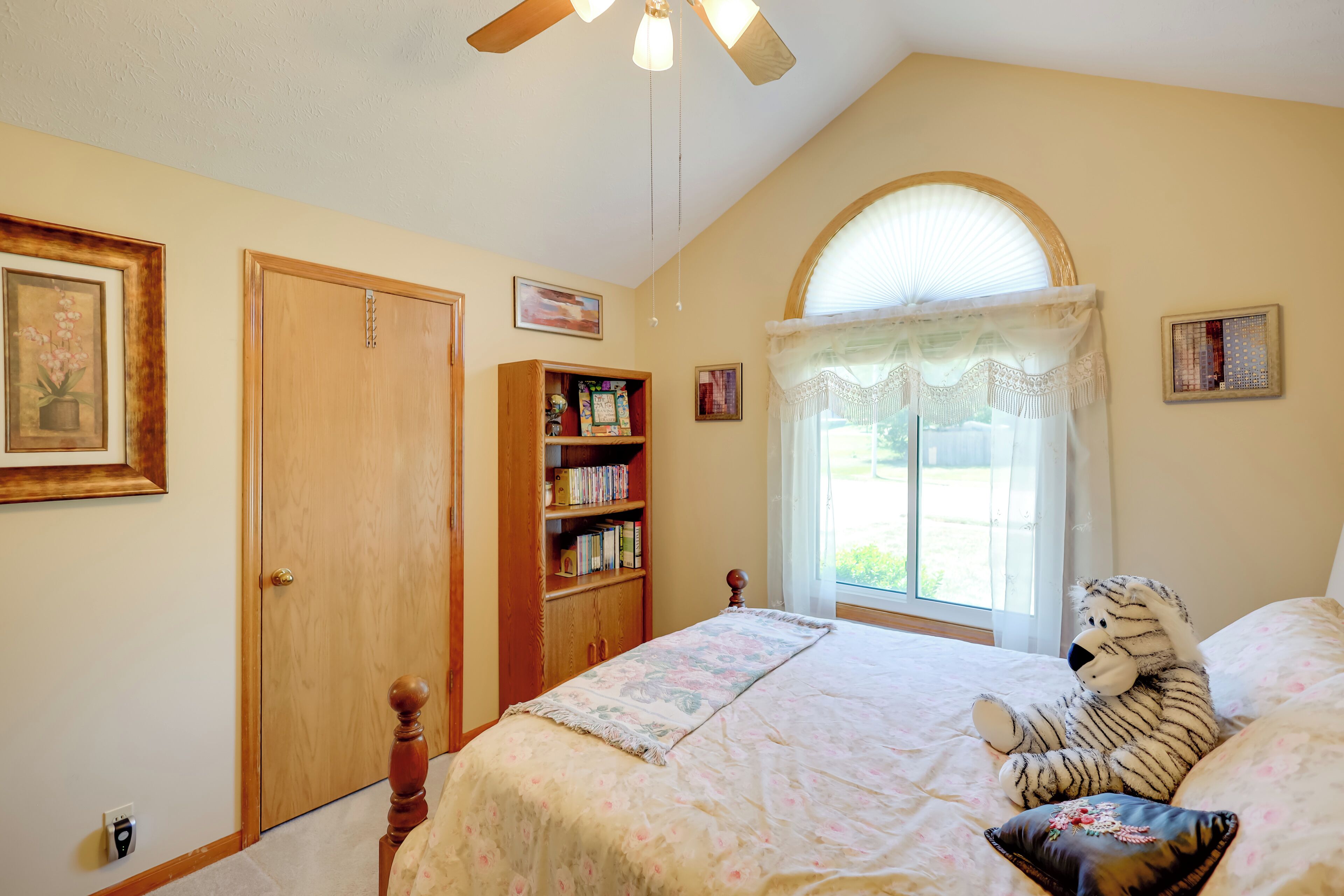 A bedroom with a bed, a wooden bookshelf, a ceiling fan, and a window with curtains.