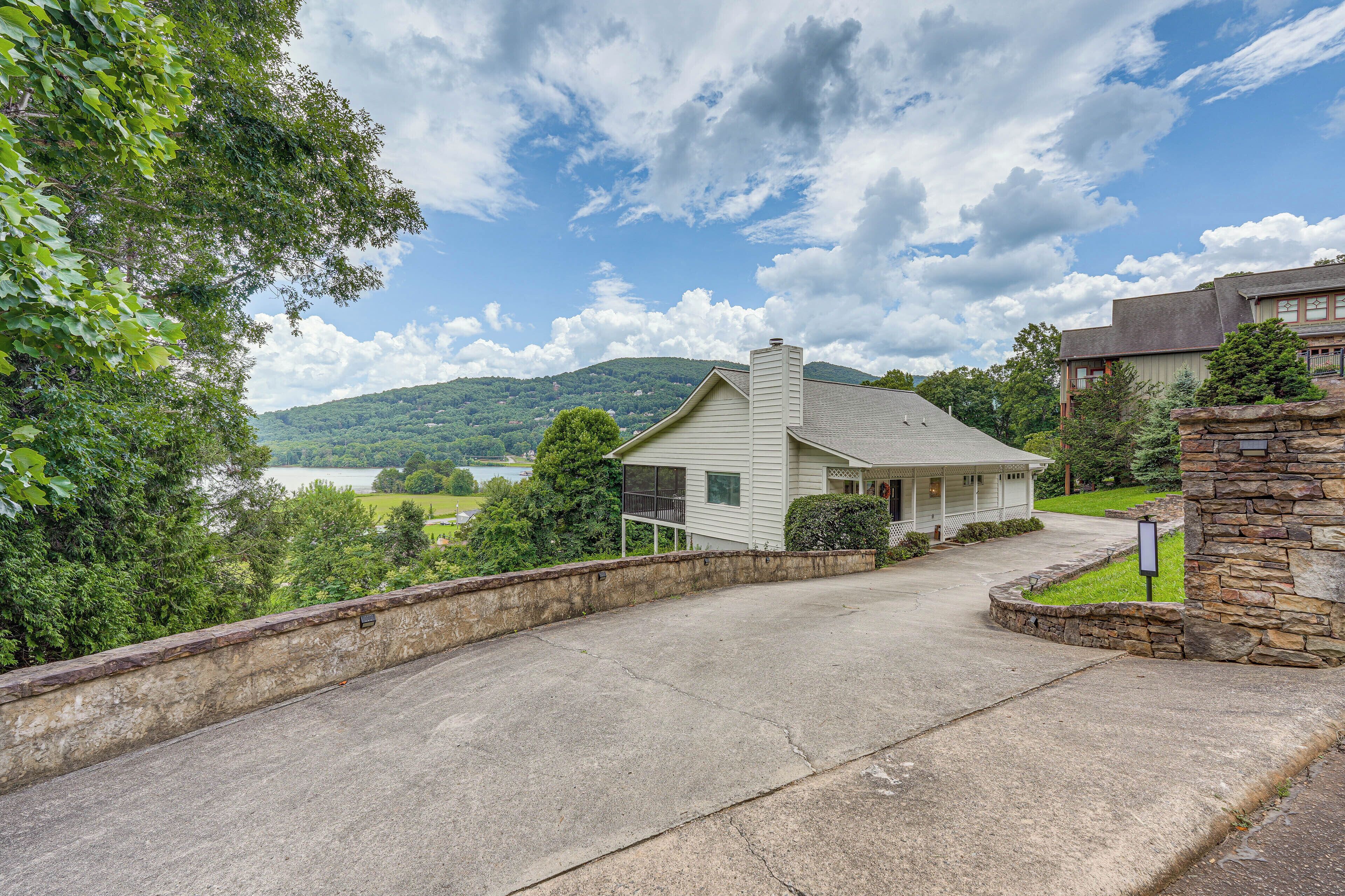 A residential street with a house featuring a stone foundation and a view of a lake and mountains in the background.