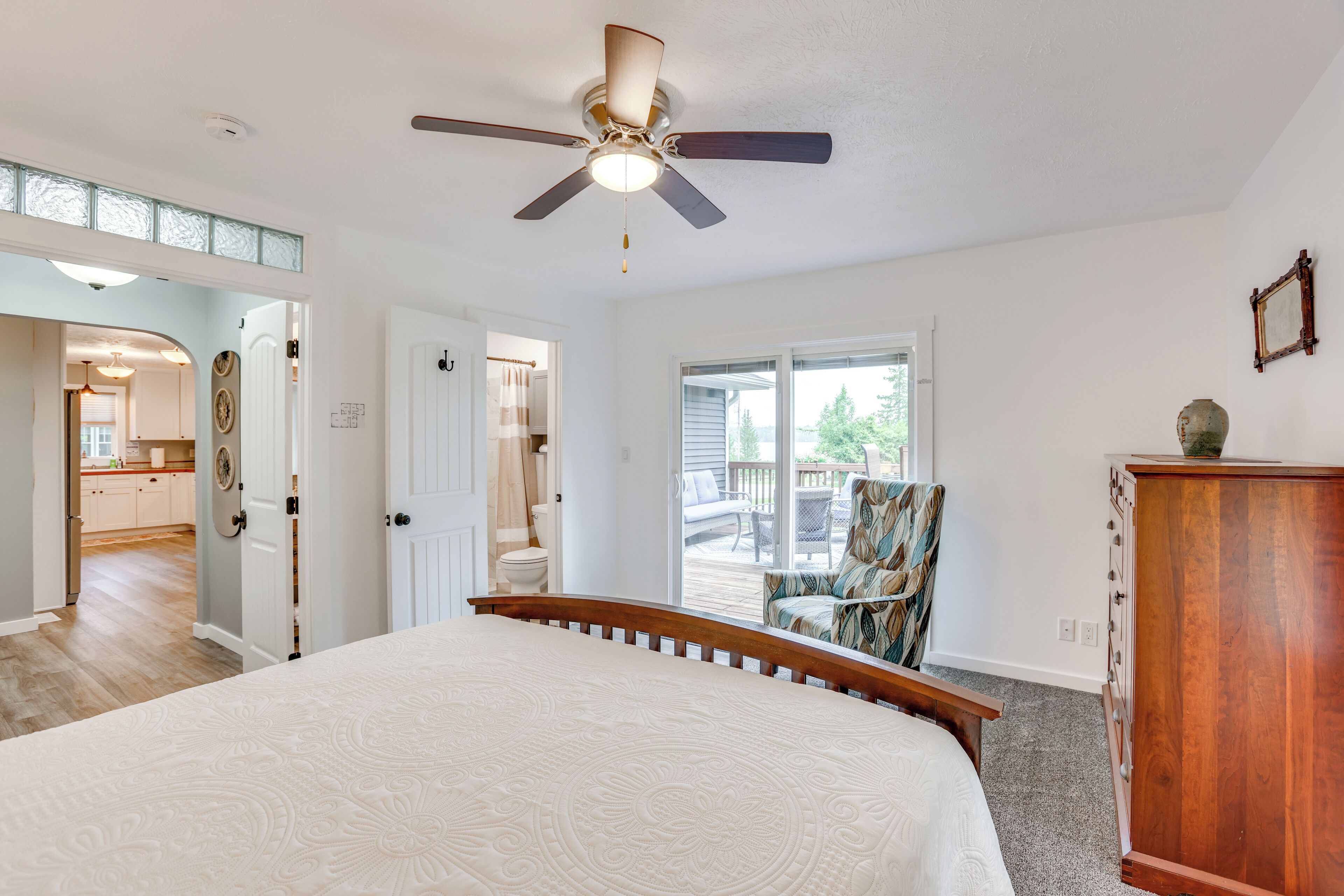A bedroom with a bed, a ceiling fan, a wooden dresser, and a view of the backyard through a sliding glass door.