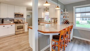 A modern kitchen with white cabinets, stainless steel appliances, a wooden island with bar stools, pendant lighting, and a large window providing a view of the outdoors. - Bring Your Boat! Little Traverse Lake Getaway (Maple City)