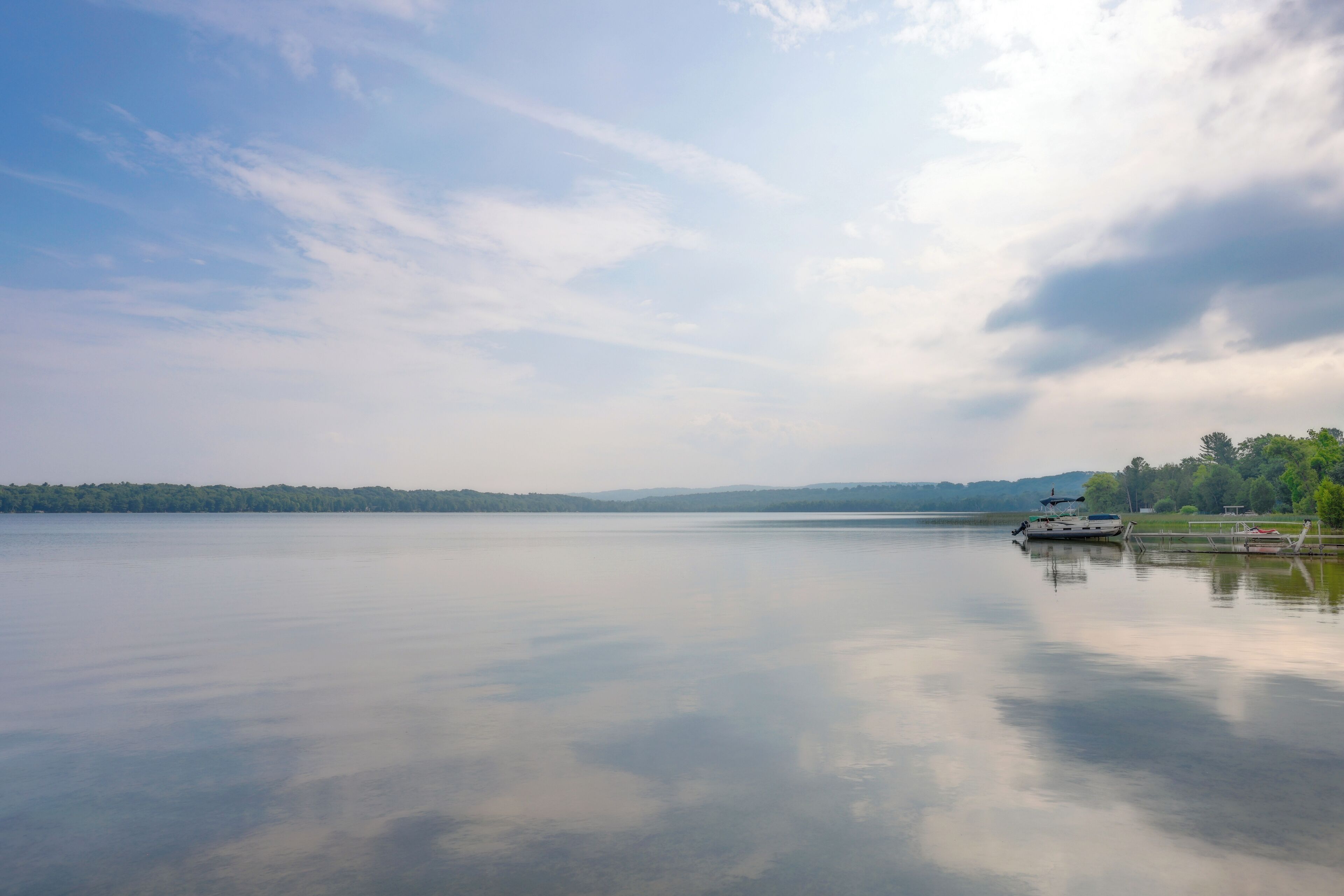Un lago tranquillo con un molo e una barca, circondato da alberi e colline lontane.