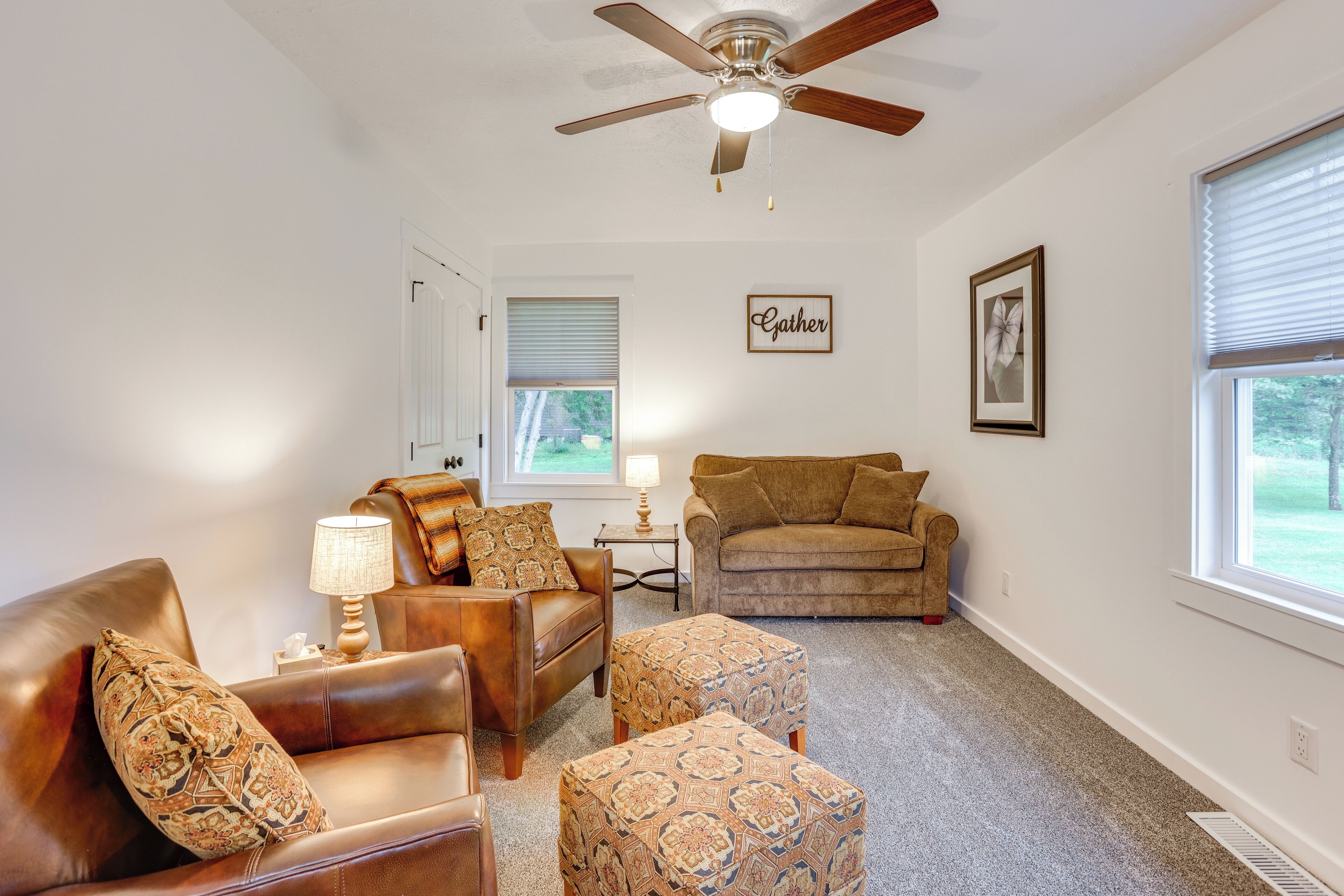 A living room with a ceiling fan, a brown sofa, patterned ottomans, a small table with a lamp, and a framed word on the wall.