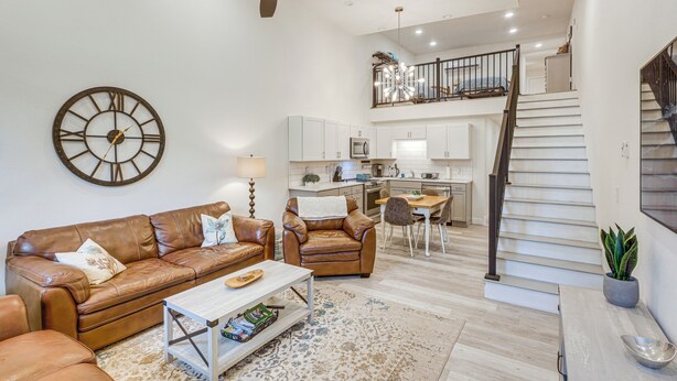 A spacious living room with a brown leather sofa, a coffee table, and a large wall clock. A staircase leads to an upper level with a kitchen and dining area. - Joliet Main Street Retreat < 30 Mi to Red Lodge (Joliet)