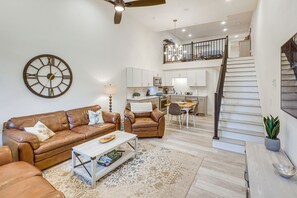 Un salon spacieux, doté d’un canapé en cuir marron, d’une table basse et d’une grande horloge murale. Un escalier mène à un niveau supérieur où se trouvent une cuisine et un coin repas.