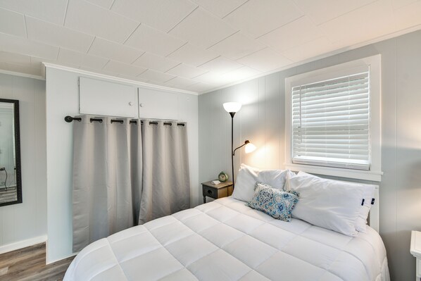 A bedroom with a bed, a nightstand, a lamp, a window with blinds, and a curtain. - Steps to Water: Houghton Lake Cottage! (Houghton Lake)