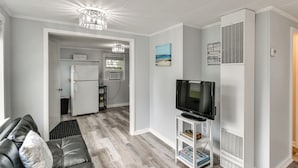 Een moderne woonkamer met een zwarte leren bank, een flatscreen televisie op een wit meubelstuk, een kroonluchter en een zichtbare keuken door een open deur.