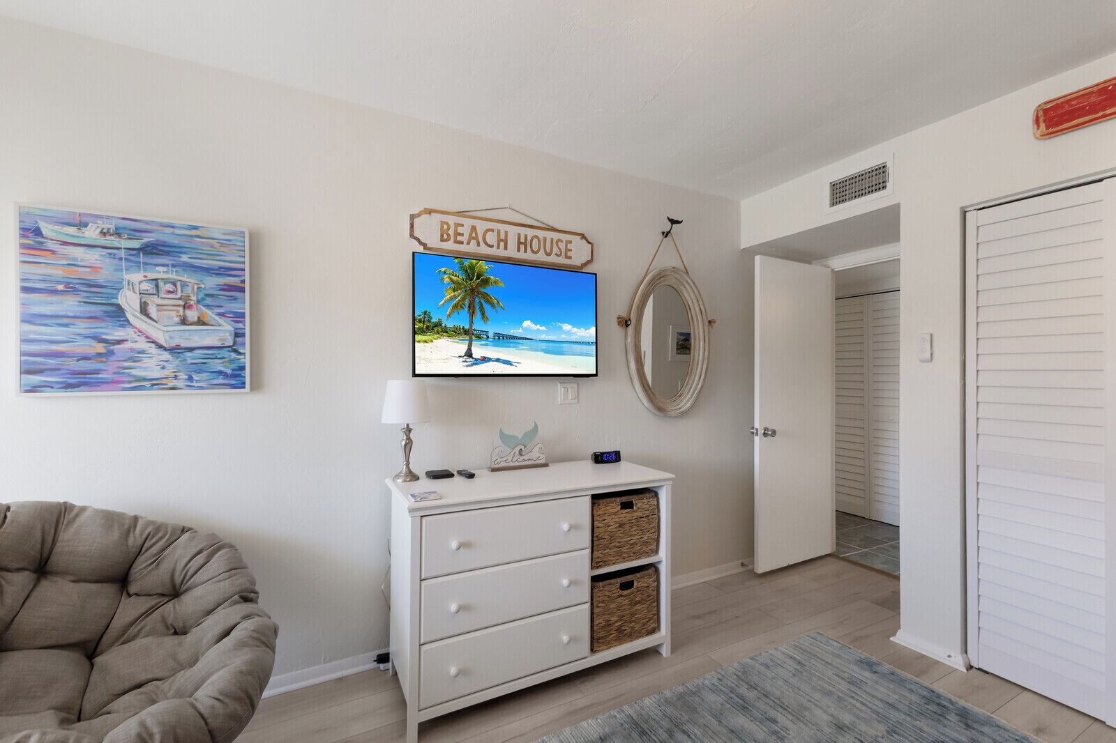 Una habitación con un aparador blanco, un televisor mostrando una escena de playa, un letrero que dice 'Beach House' y un cuadro de un bote.