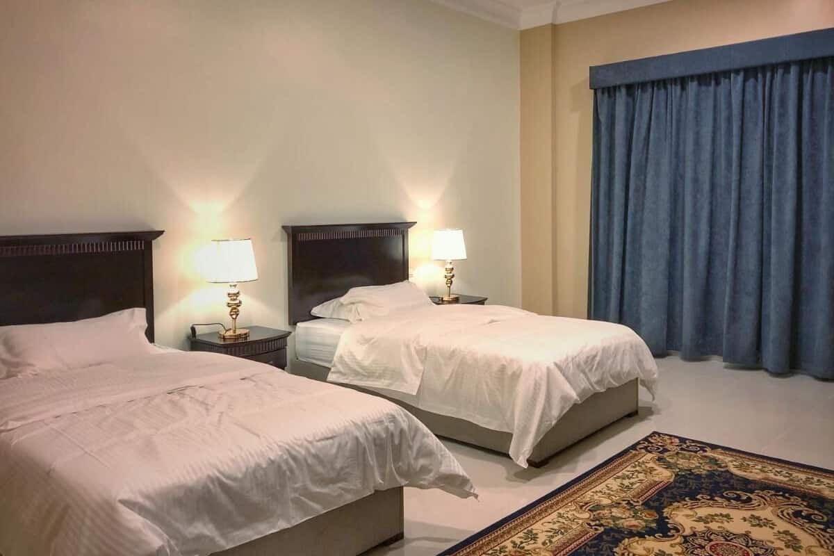 Une chambre d’hôtel avec deux lits, une table de chevet avec des lampes, une grande fenêtre avec des rideaux bleus et un tapis à motifs.