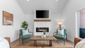 Een moderne woonkamer met een open haard, twee groene fauteuils, een salontafel en een flatscreen televisie die boven de haard is bevestigd.