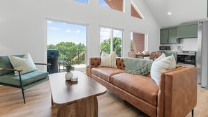Een ruime woonkamer met een bruine leren bank, een houten salontafel en een groene fauteuil. Achtergrond toont een keuken met groene kastjes en een eethoek met een tafel en stoelen.