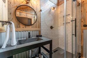 A bathroom with a wooden wall, a round mirror, a sink, and a glass shower enclosure.