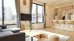 Una sala de estar moderna con un sofá, una mesa de centro y un televisor. Cuenta con una cocina con mostrador y taburetes. Amplias ventanas ofrecen vistas de la ciudad.