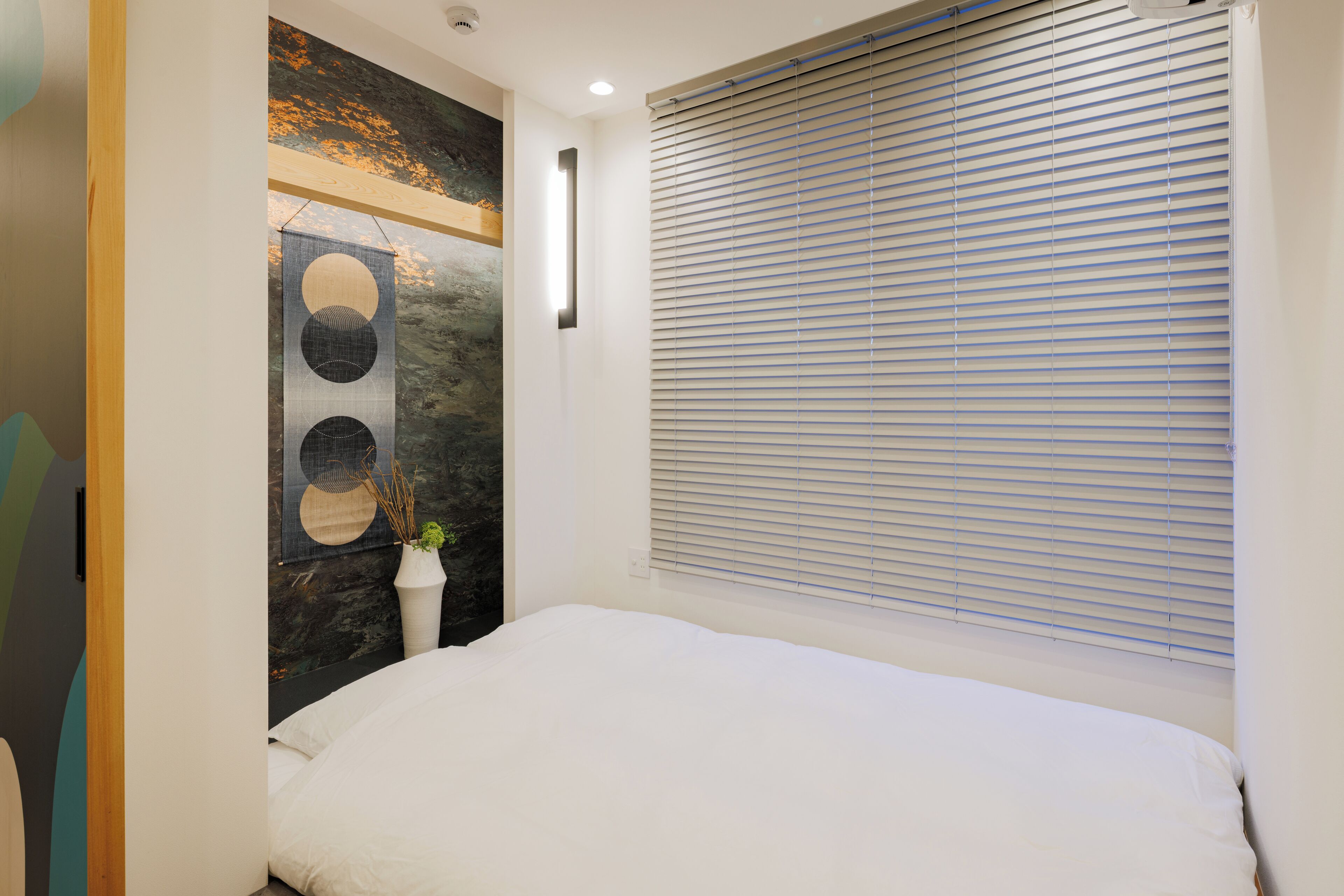 A bedroom with a bed, a wall-mounted artwork, a window with blinds, and a small bedside table with a vase.