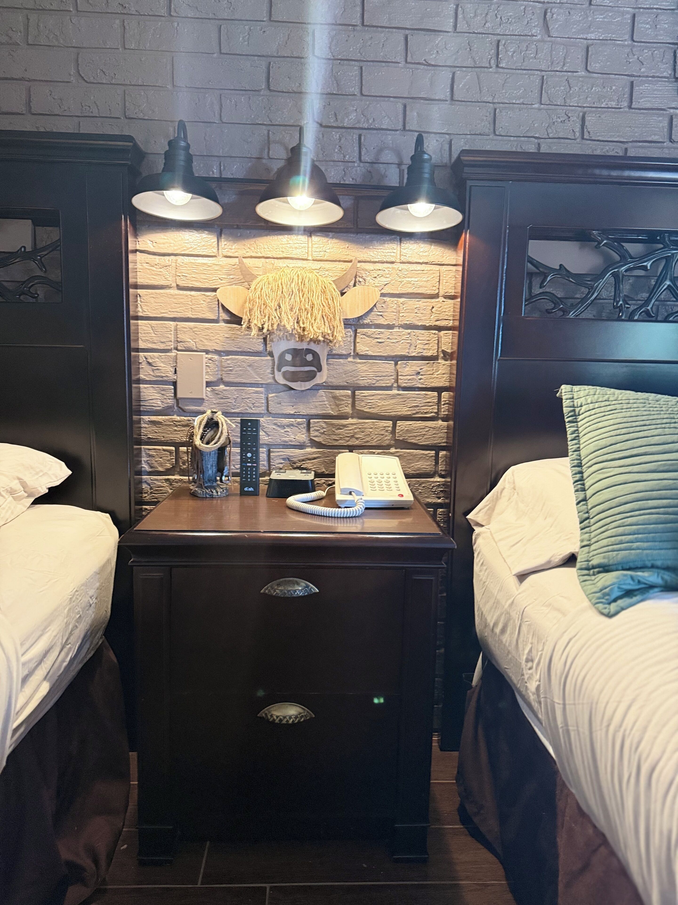 A hotel room with a brick wall, a headboard with a hornet design, two bedside tables with lamps, a telephone, and a decorative hornet head.