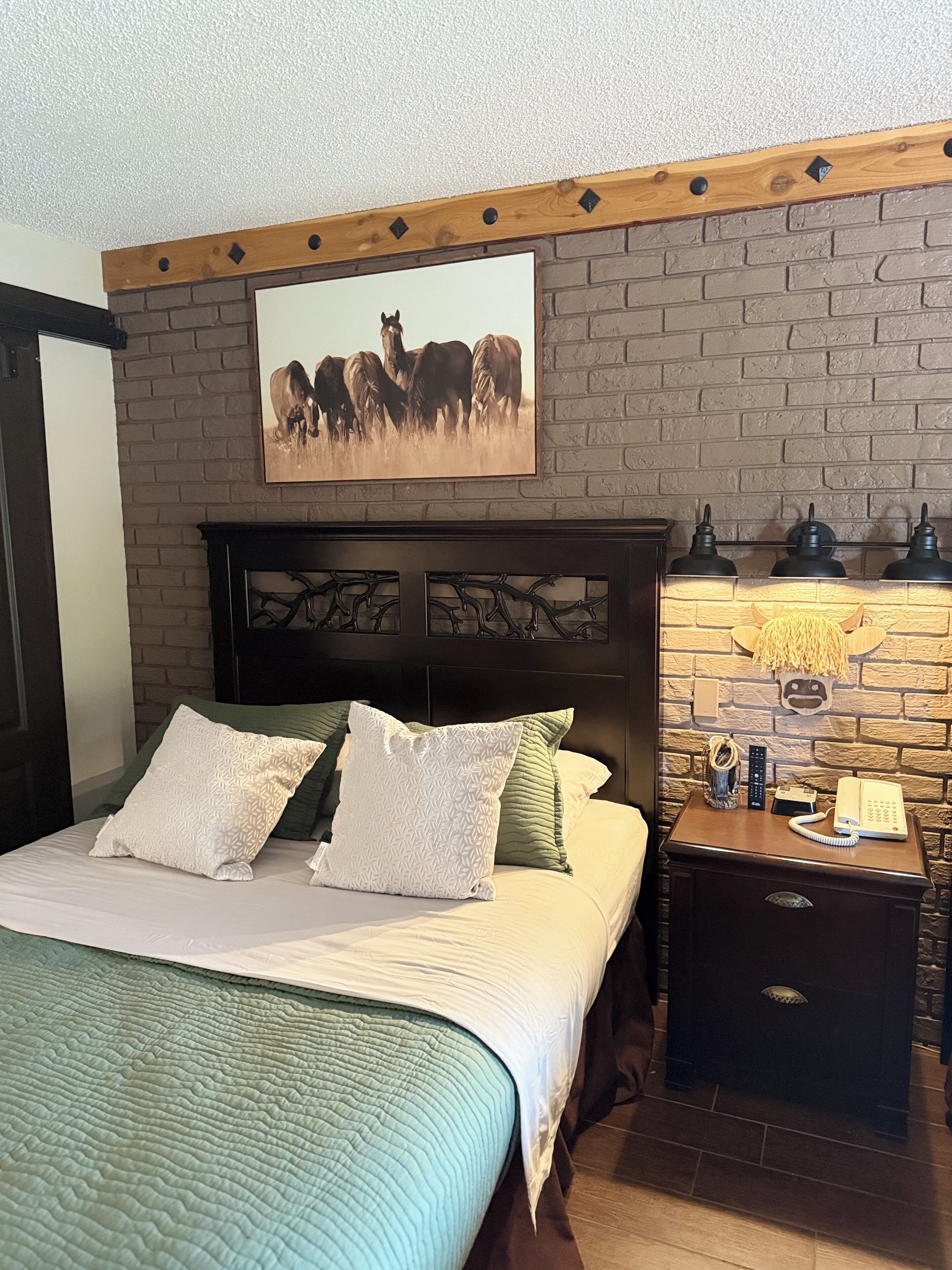 A bedroom with a brick wall, a wooden headboard, a bed with white and green pillows, a nightstand with a lamp, a phone, and a remote control.