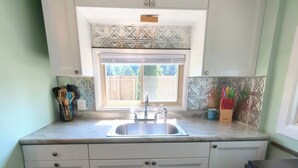 A compact kitchen with white cabinets, a stainless steel sink, and a window with a view of a wooden deck. - Cozy Haven - Little Lakefront Home (Little Lake)
