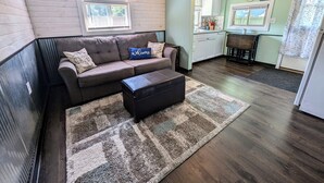 A cozy living room with a brown sofa, a coffee table, and a patterned rug. A kitchen with white cabinets and a window is visible in the background. - Cozy Haven - Little Lakefront Home (Little Lake)