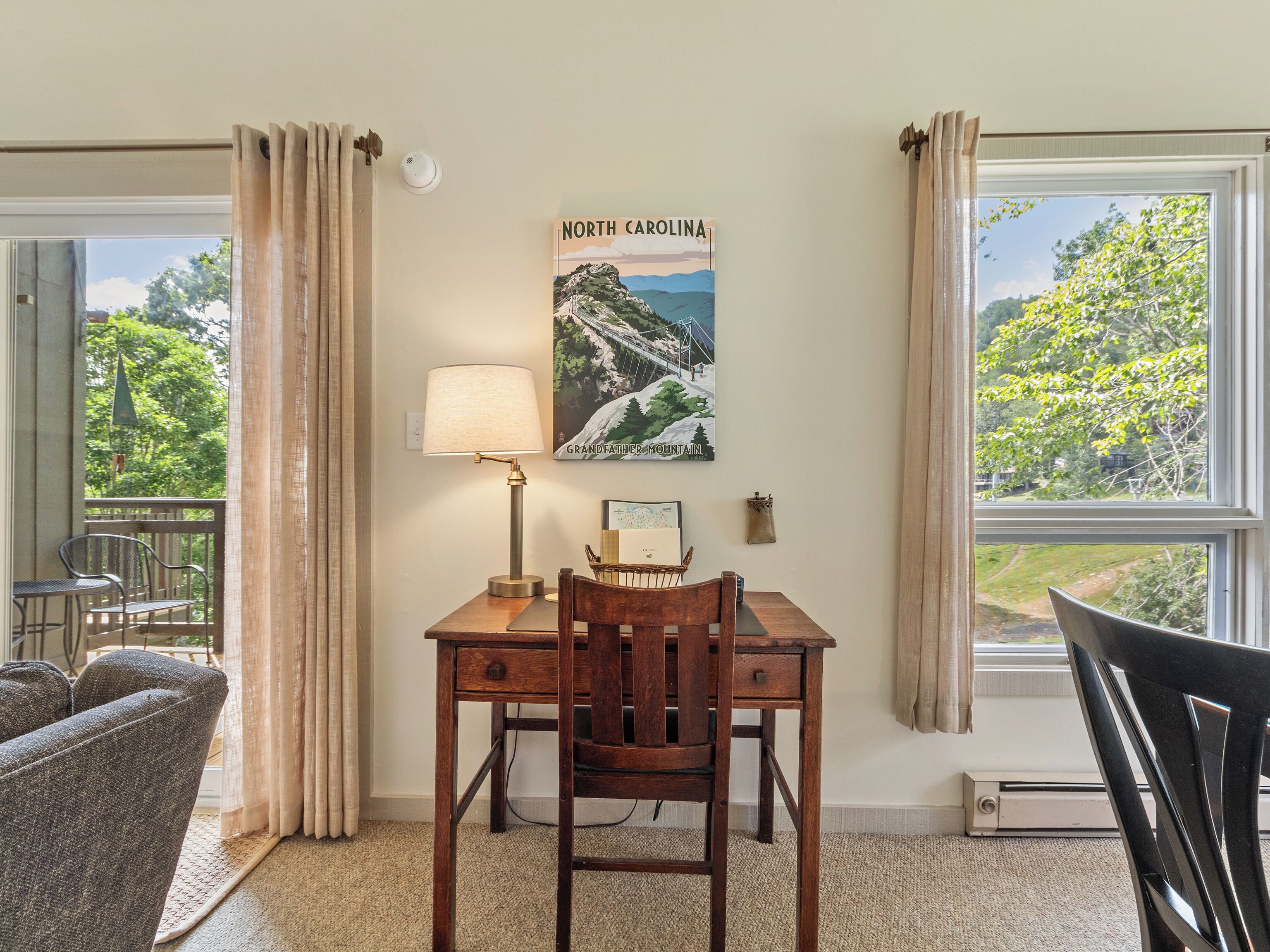 A room with a dining table, chairs, a lamp, a poster of North Carolina's Grandfather Mountain, and a view of greenery through the windows.