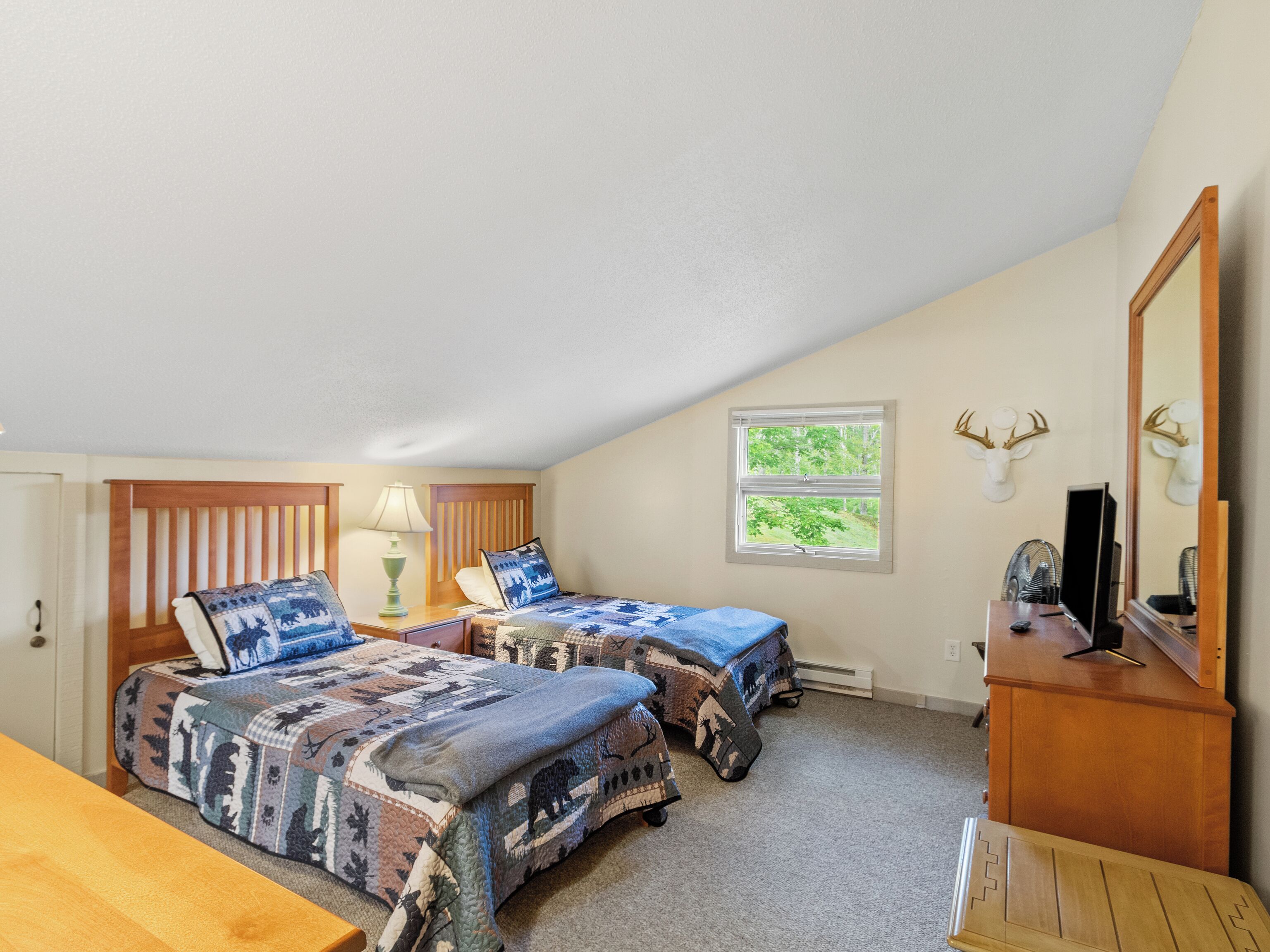 A bedroom with two beds, a dresser, a mirror, and a window with a view of trees.
