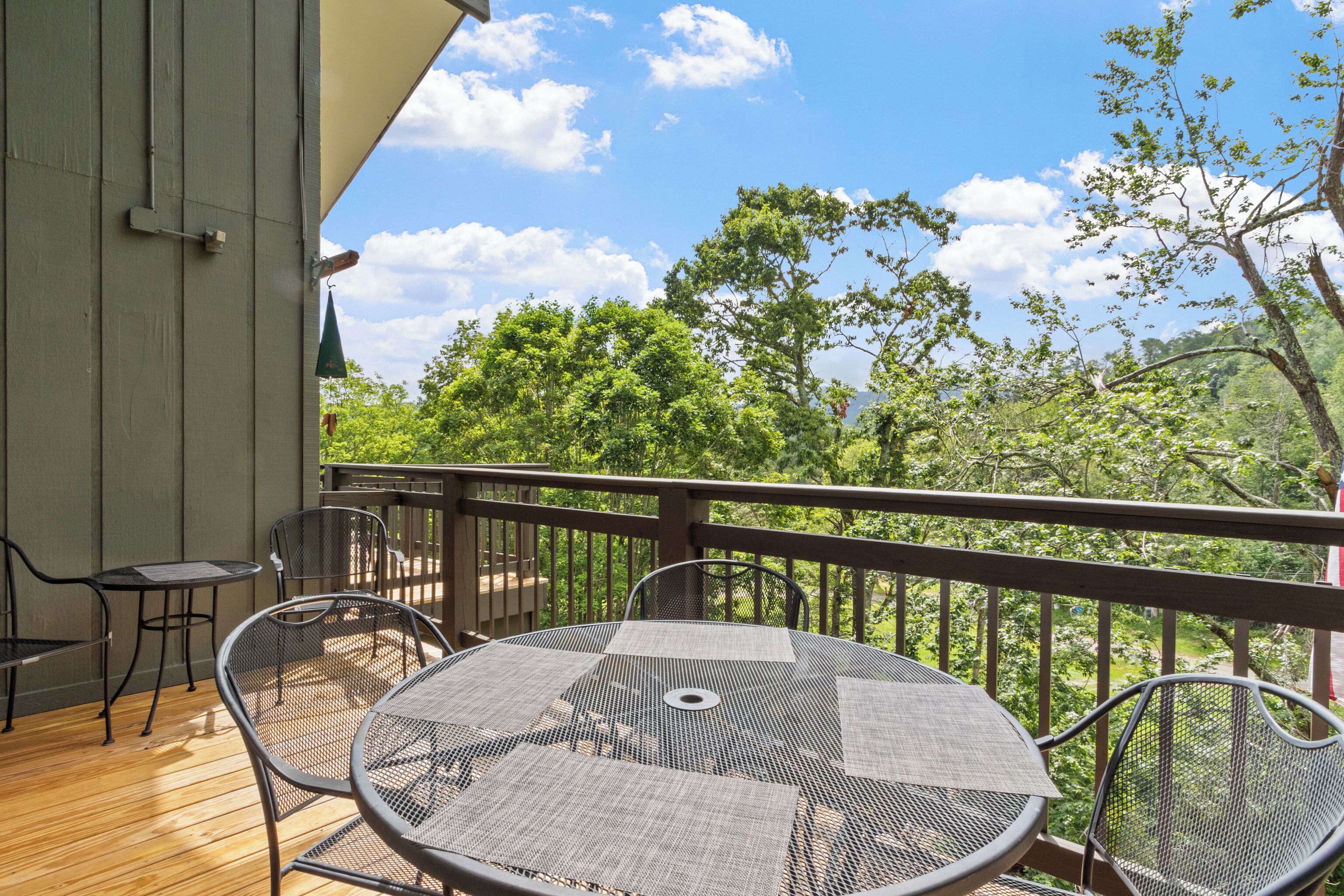 A covered outdoor area with a round table and chairs, a wooden deck, and a view of trees and sky.