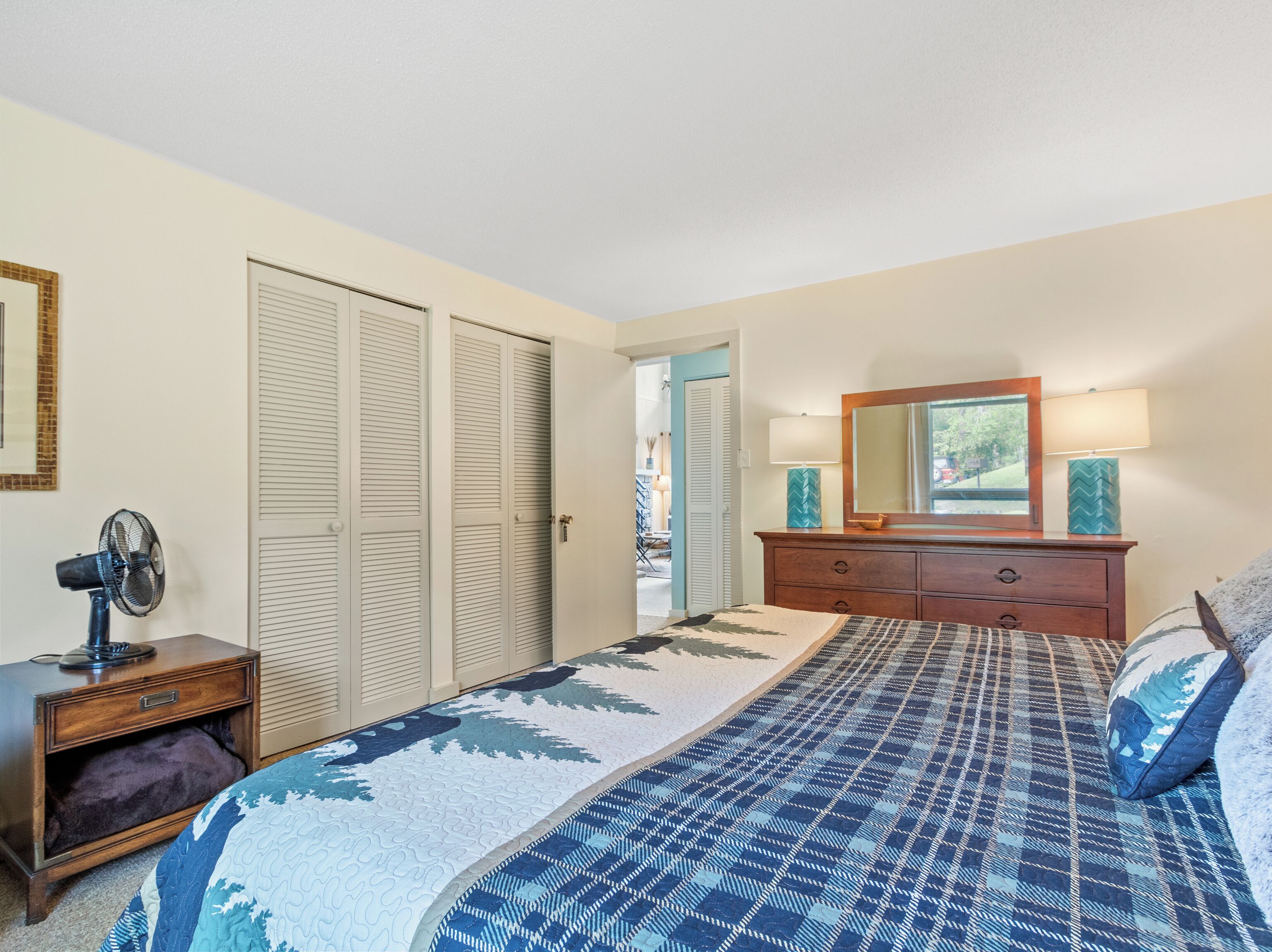 A bedroom with a bed, a dresser, a mirror, a fan, and a door leading to another room.