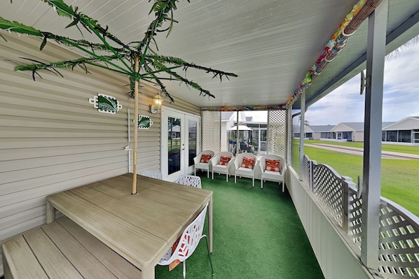 A covered patio with a wooden table and chairs, a green artificial turf floor, and a view of houses through a lattice fence.