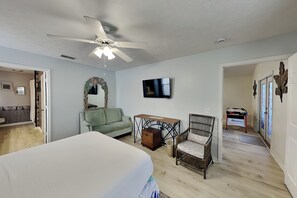 A bedroom with a bed, a ceiling fan, a TV, a wicker chair, a small table, and a wooden chest.