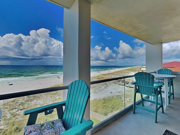 Un balcon avec des chaises turquoise et une table donnant sur une plage, sous un ciel dégagé.
