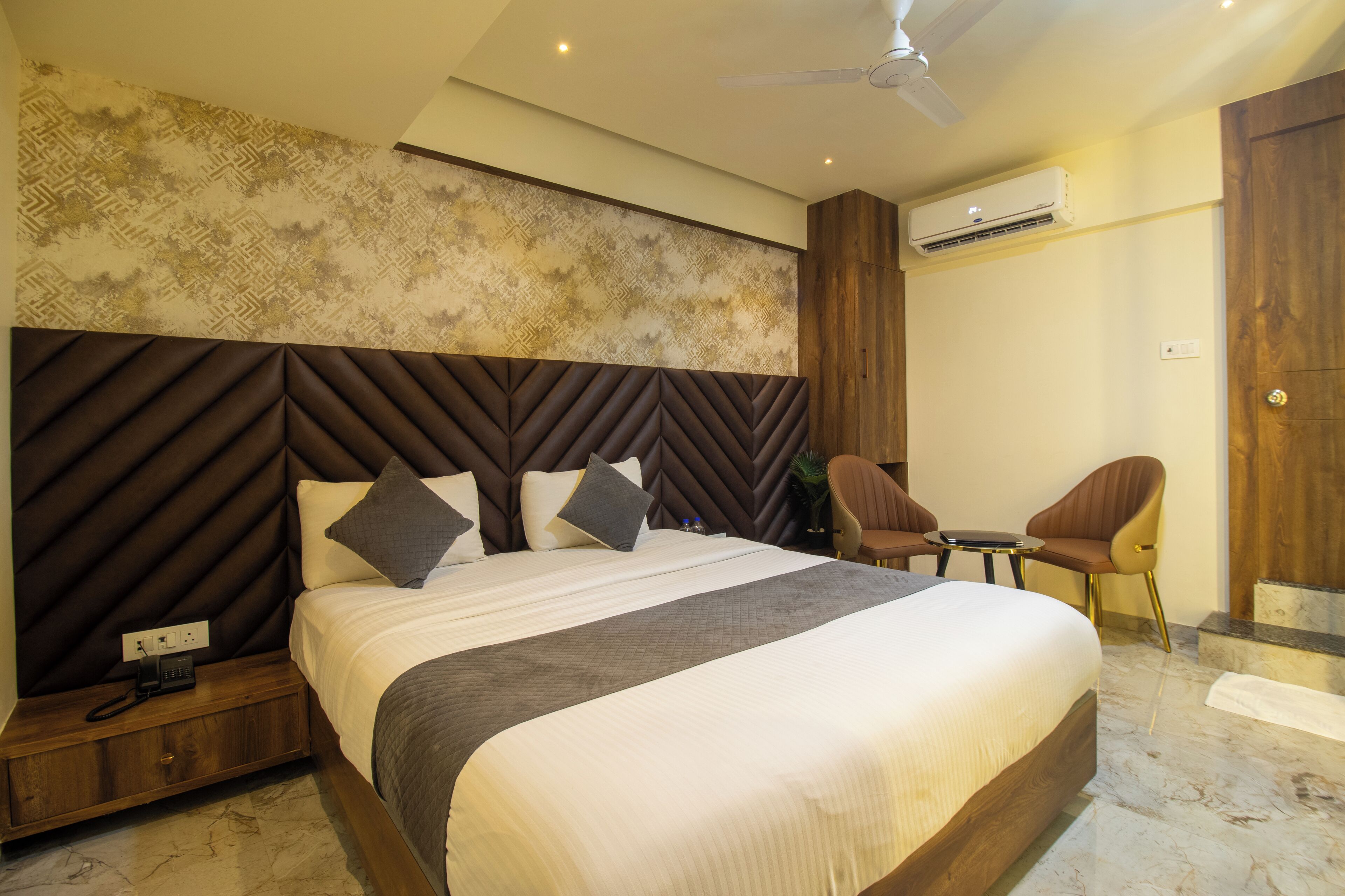 A modern hotel room with a large bed, a wooden headboard, a small table, two chairs, and a ceiling fan.