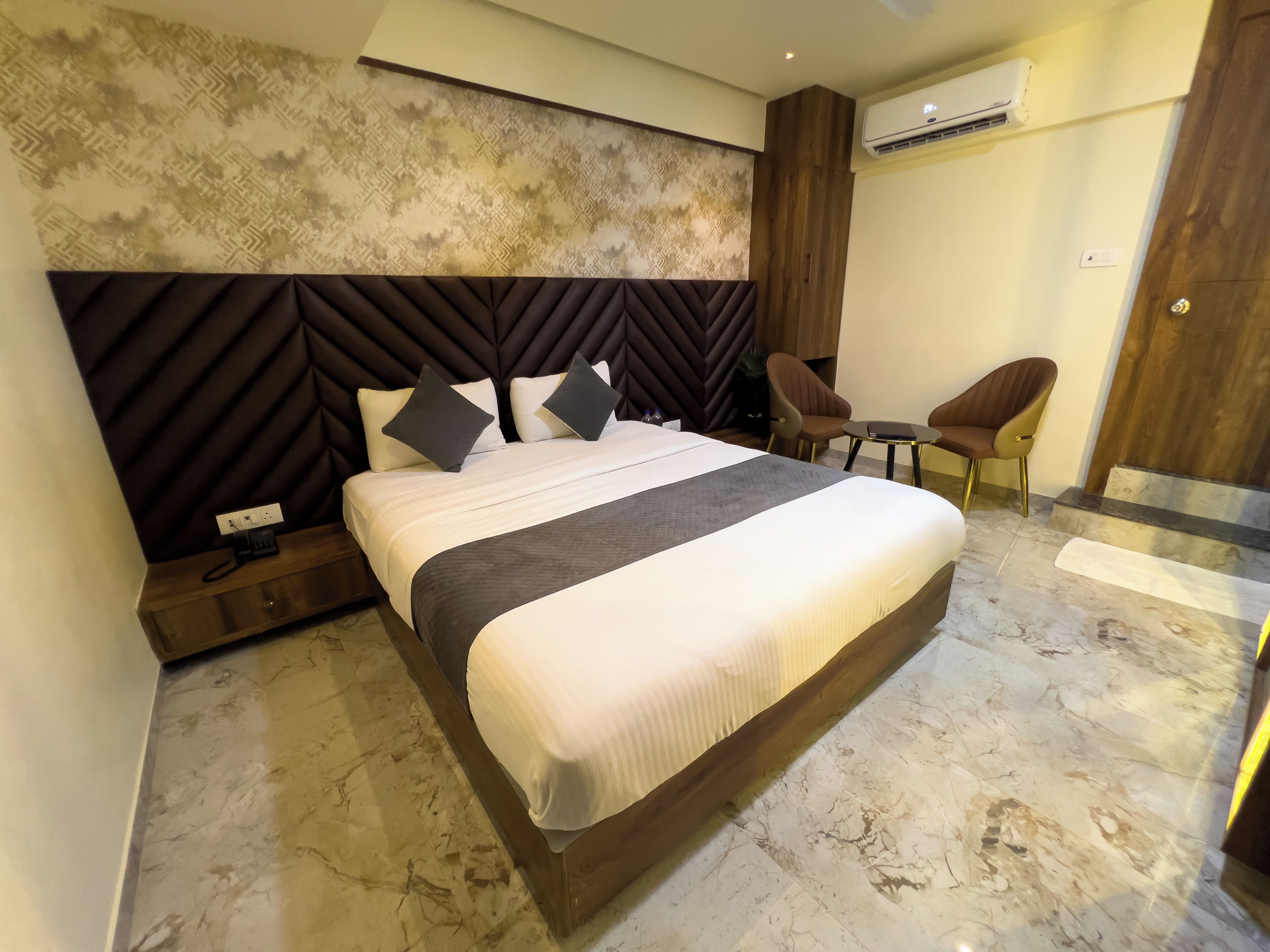 A modern hotel room with a large bed, a headboard with a geometric pattern, a small table, two chairs, and an air conditioning unit on the wall.