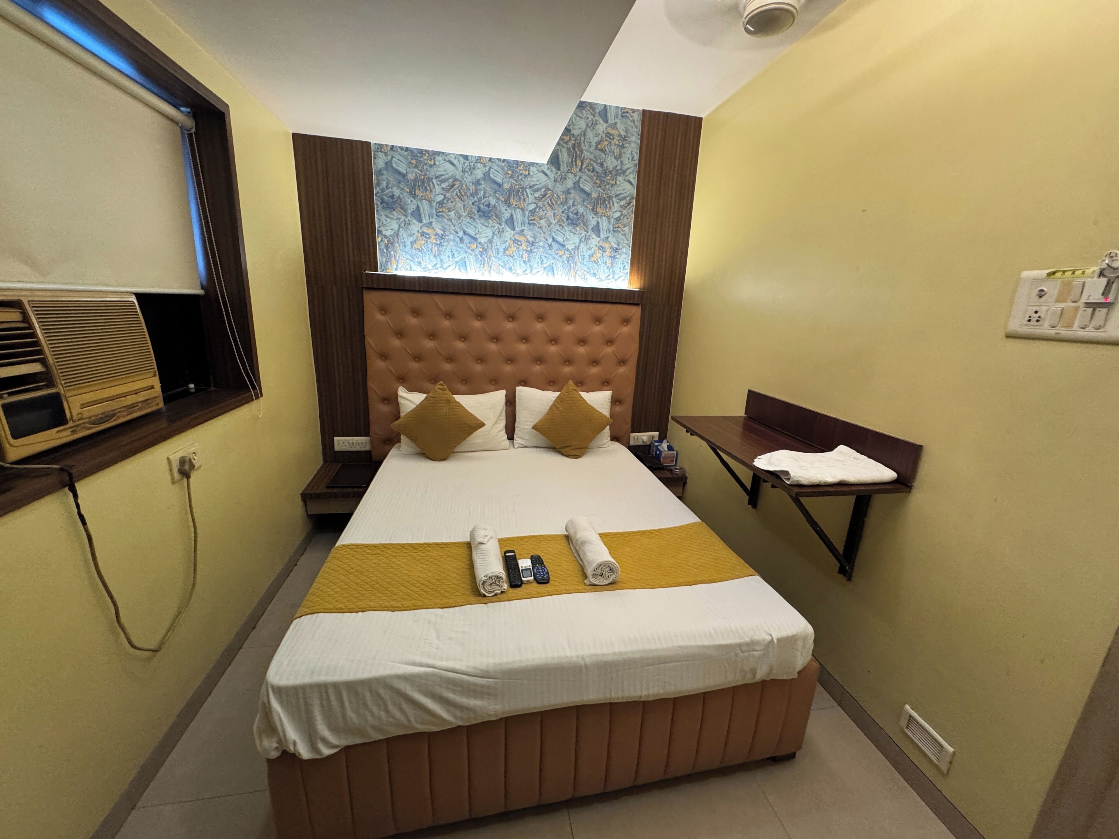A hotel room with a large bed, a bedside table, a small desk, a window with blinds, and a wall-mounted air conditioning unit.