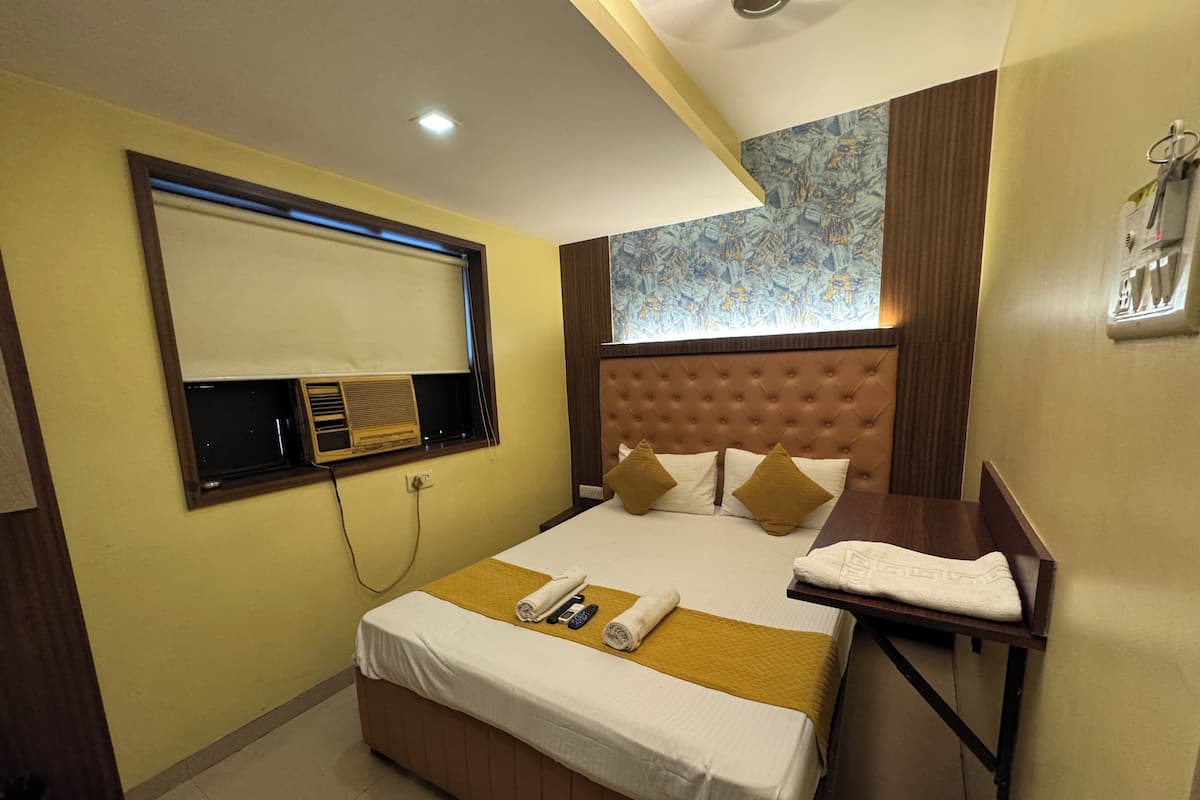 A hotel room with a bed, a bedside table, a window with blinds, a wall-mounted air conditioning unit, and a ceiling light.