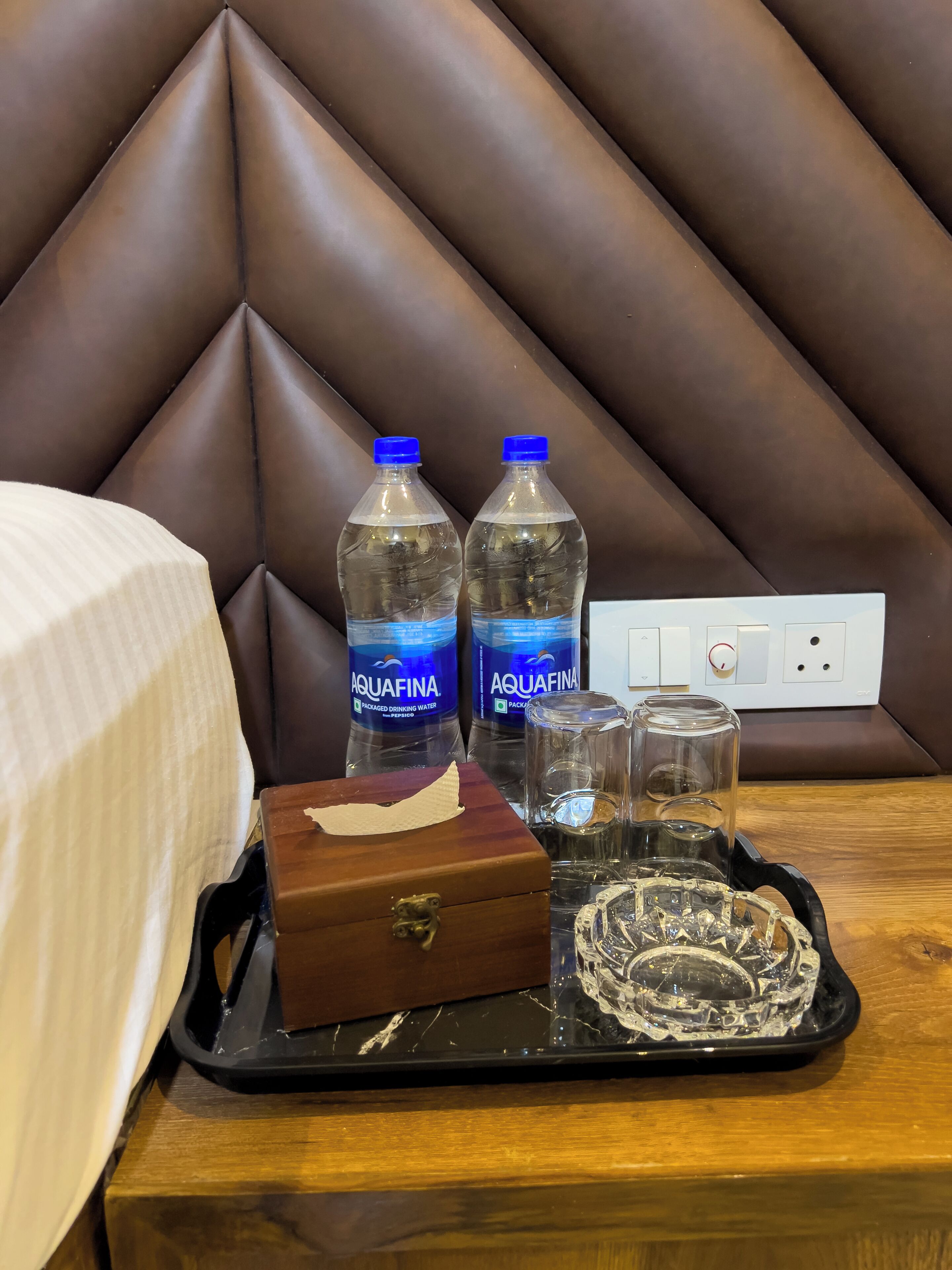 A hotel room with a brown headboard, a wooden bedside table, two Aquafina water bottles, a tissue box, and a tray with a glass and a crystal bowl.
