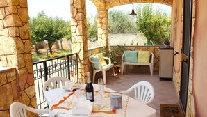 A patio with a table set for two, white chairs, a bottle of wine, and a glass. There are potted plants and a small table with a cushion. The patio is enclosed by stone walls and has a view of trees and a fence in the background. - RIVER VIEW CHARMING VILLA IN AMAZING ALGHERO WITH AC (Alghero(SS))