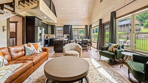 A spacious living area with a high ceiling, wooden beams, a central island, and large windows offering views of the outdoors. - New! 3BR w/Hot Tub*Arcade*5 Min to Pigeon Forge (Pigeon Forge)