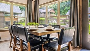 A dining area with a table set for four, surrounded by chairs, large windows with curtains, and a view of a wooden deck and buildings. - New! 3BR w/Hot Tub*Arcade*5 Min to Pigeon Forge (Pigeon Forge)