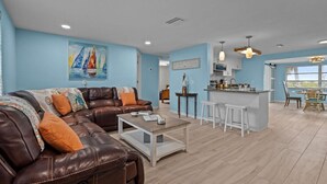 A spacious living room with a brown leather sofa, a coffee table, and a painting of a sailboat. A kitchen area with a bar and stools is visible in the background. - Waterfront! 2BR w/Firepit & Grill! (Hudson)