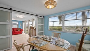 A dining area with a round table set for four, surrounded by wooden chairs with green cushions. A red sofa is visible in the background, and the room has large windows with blinds. - Waterfront! 2BR w/Firepit & Grill! (Hudson)