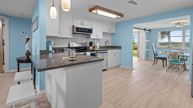 A modern kitchen with white cabinets, granite countertops, and stainless steel appliances. There is a dining area with a table and chairs, and a view of the outdoors through large windows. - Waterfront! 2BR w/Firepit & Grill! (Hudson)