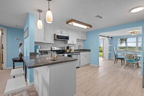 A modern kitchen with white cabinets, granite countertops, and stainless steel appliances. There is a dining area with a table and chairs, and a view of the outdoors through large windows.
